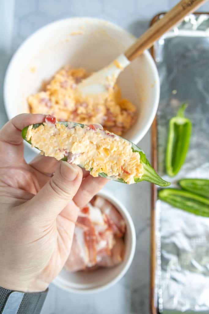 A hand holds a halved jalapeño pepper stuffed with a creamy cheese mixture; bowls of filling and bacon, a baking sheet, and more jalapeños are visible in the background.