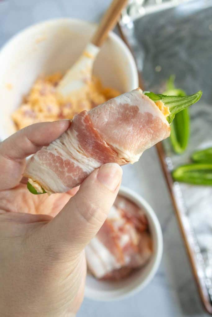 A hand holds a jalapeño stuffed with cheese mixture and wrapped in bacon, with a mixing bowl and prepared ingredients in the background.