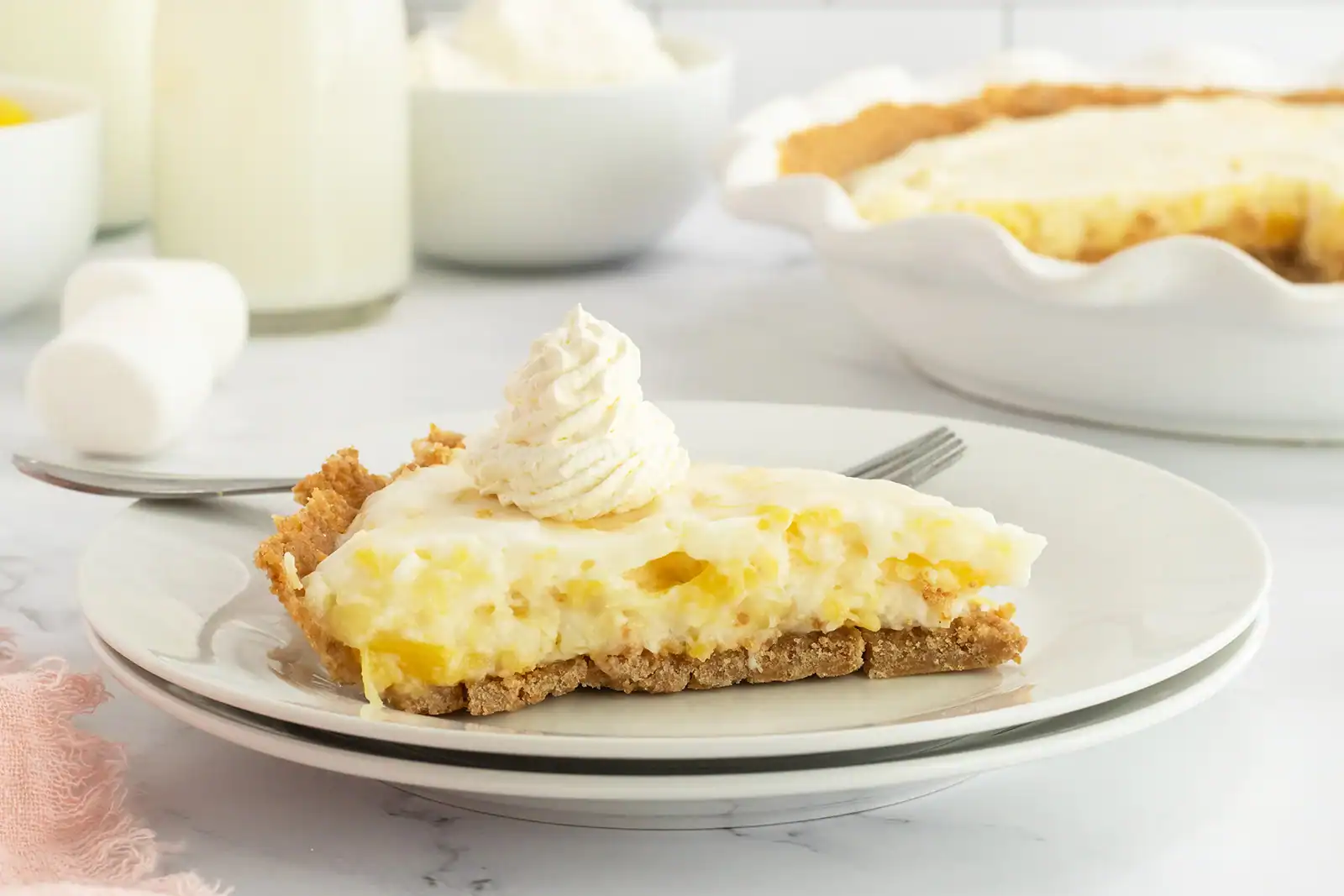 A slice of creamy pie with a graham cracker crust and whipped topping on a plate, with a fork beside it and a whole pie in the background.