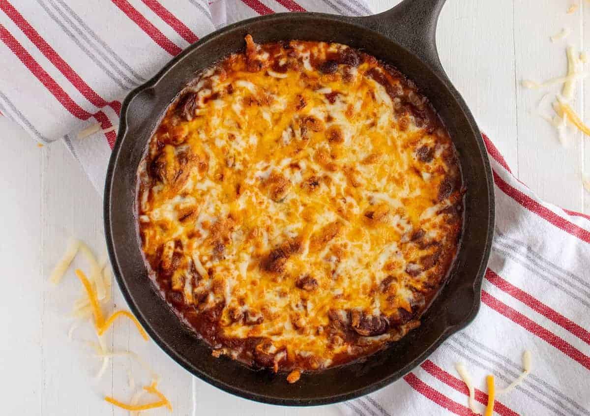 A cast iron skillet filled with a baked cheesy casserole, topped with melted cheese, sits on a striped kitchen towel on a white surface.