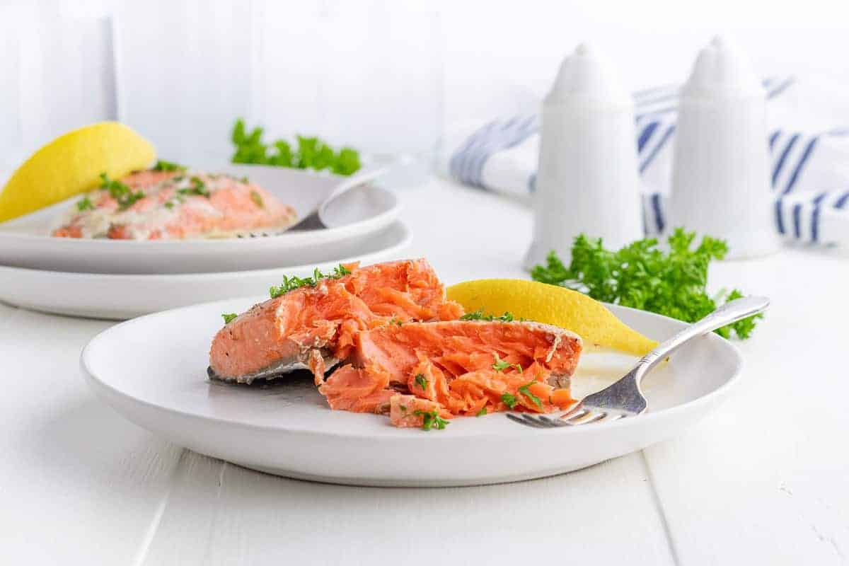 A plate of cooked salmon garnished with parsley, served with a lemon wedge and a fork on a white table; another plate is visible in the background.