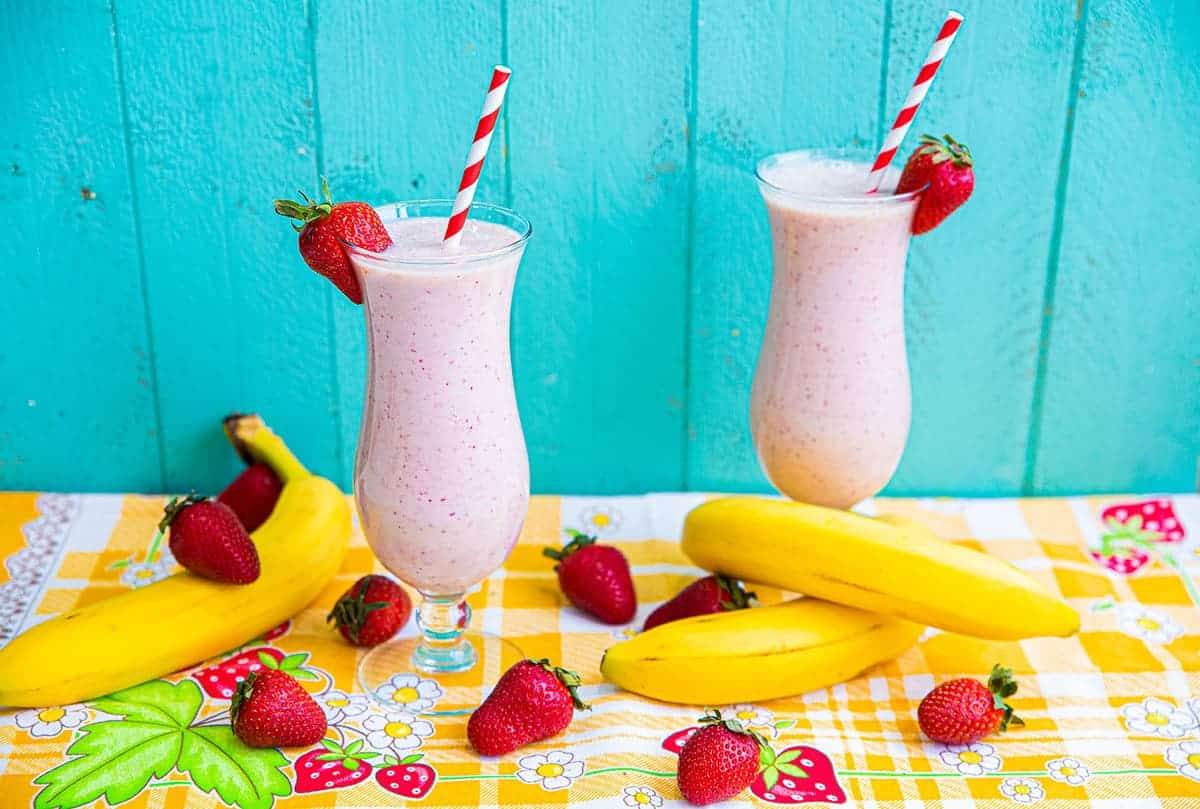 Two glasses of pink strawberry banana smoothies with red striped straws, garnished with strawberries, surrounded by bananas and strawberries on a colorful tablecloth against a blue wooden wall.