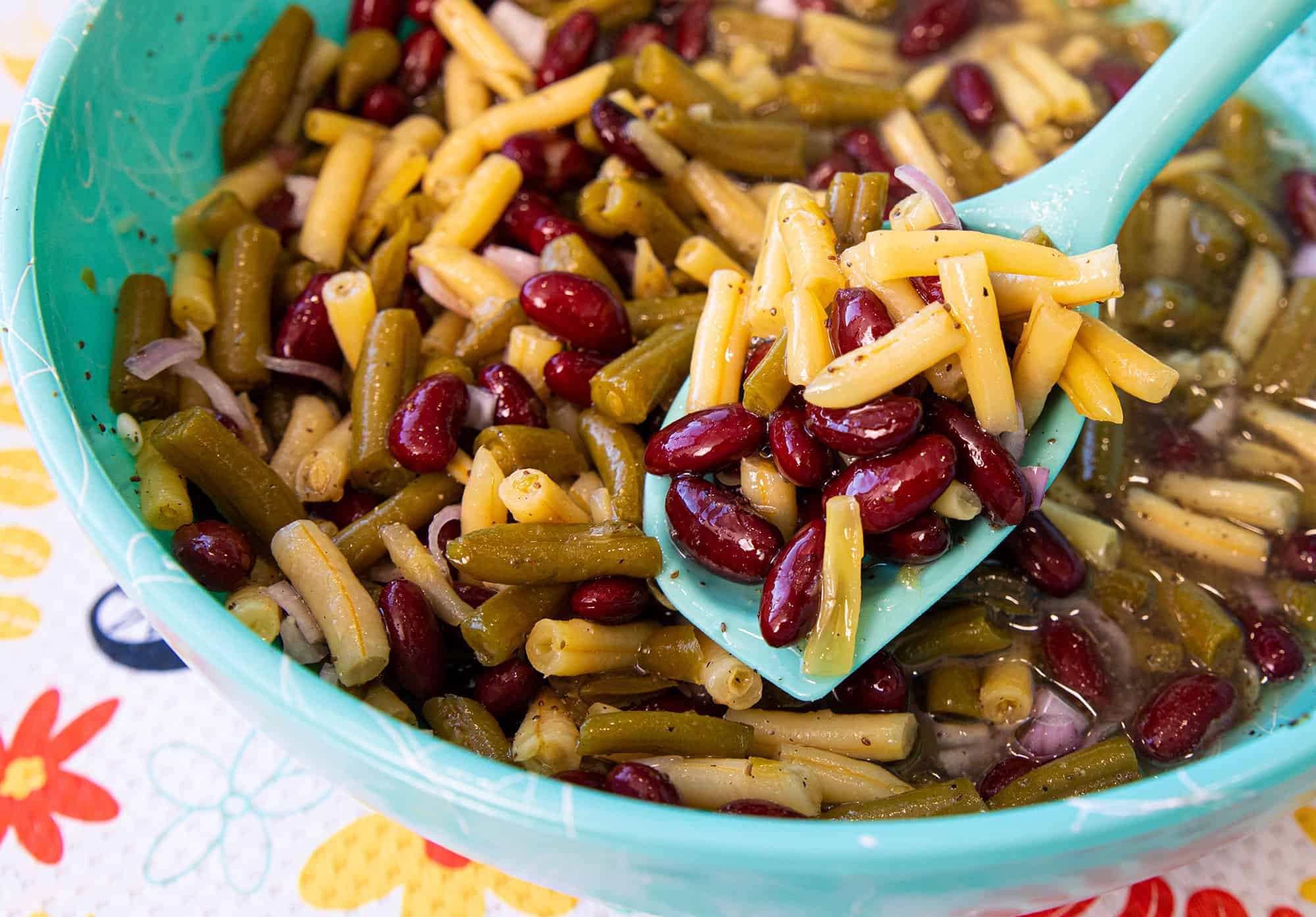 A close-up of a bowl filled with three bean salad, including kidney beans, wax beans, green beans, and chopped onions, with a spoon scooping some out.