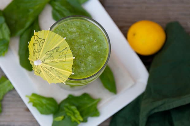 A glass of green smoothie with a yellow paper umbrella on top, surrounded by green leaves and a whole yellow lemon on a white plate.