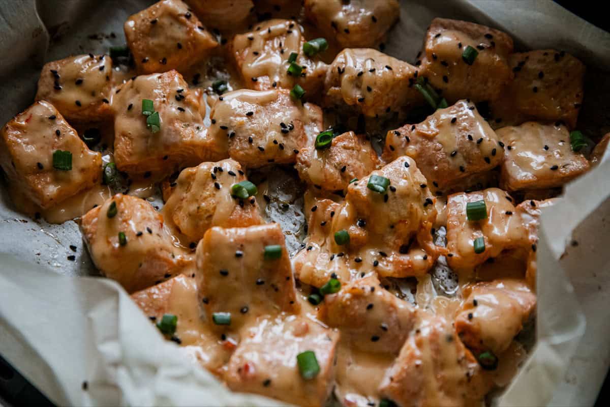 Baked salmon cubes topped with creamy sauce, black sesame seeds, and chopped green onions on parchment paper.