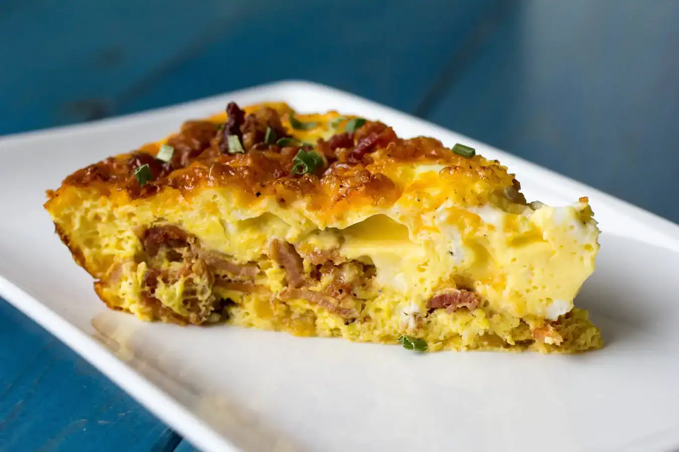 A slice of breakfast casserole with eggs, cheese, bacon, and chives on a white plate, set on a blue surface.