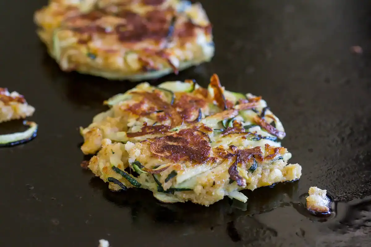 A zucchini fritter cooking on a dark, oiled skillet, with visible browning and crispy edges.