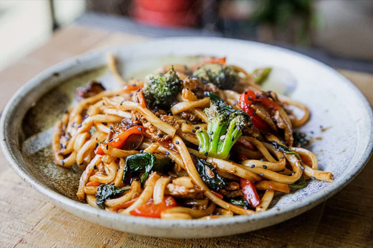A plate of stir-fried udon noodles with broccoli, red bell peppers, spinach, and a savory sauce, served on a ceramic dish.