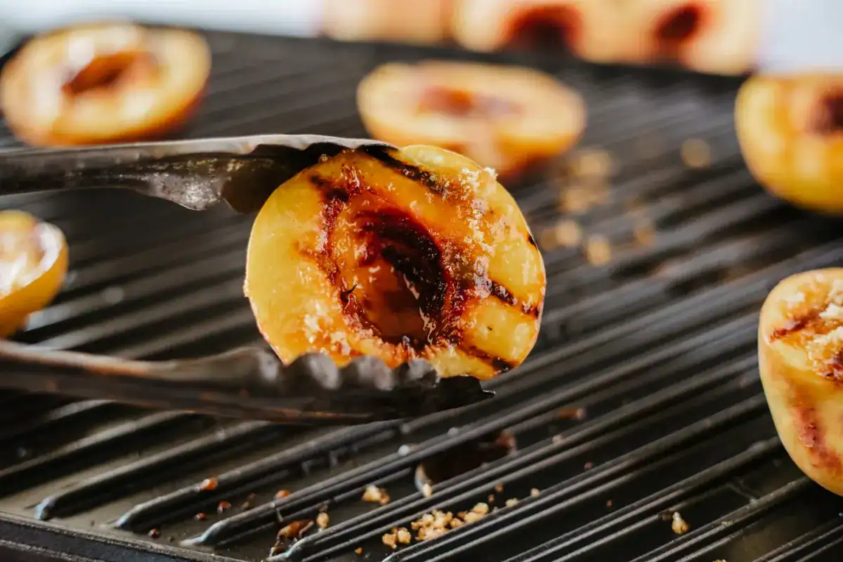 A close-up of metal tongs holding a grilled peach half over a grill with other grilled peach halves in the background.