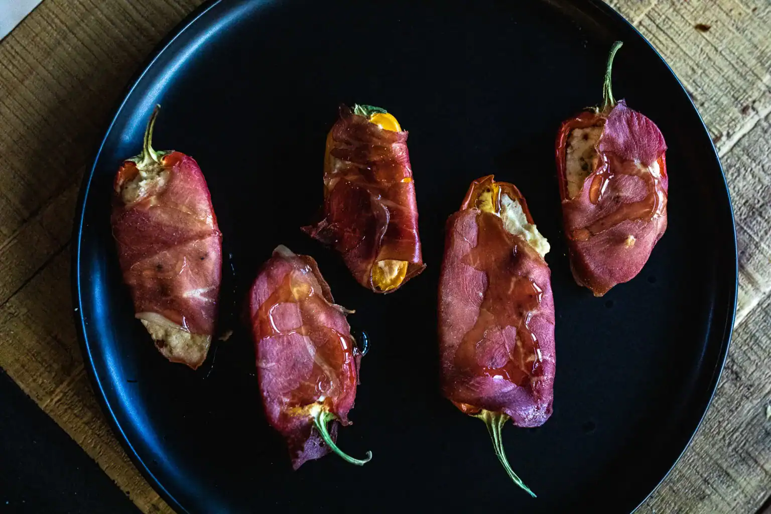 Five stuffed jalapeños wrapped in prosciutto are arranged on a black plate, set on a wooden surface.