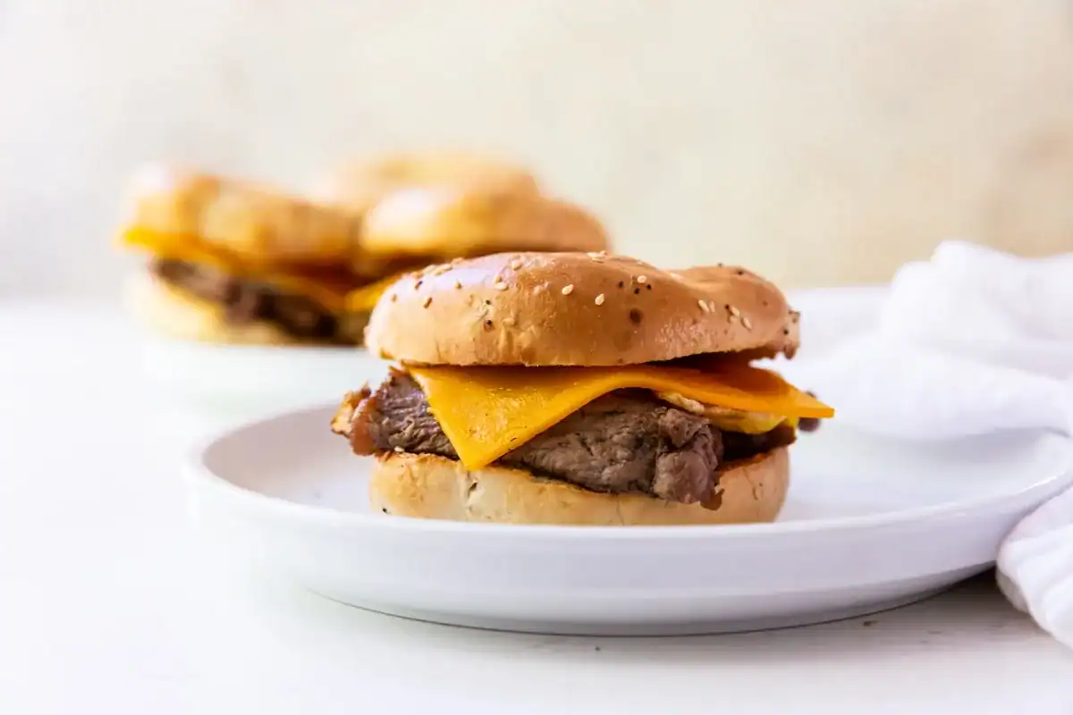 A sandwich with sliced beef and cheddar cheese on a seeded bun sits on a white plate, with another sandwich blurred in the background.