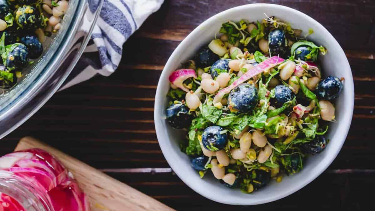 A bowl of salad containing blueberries, white beans, leafy greens, and sliced red onions on a dark wooden table.