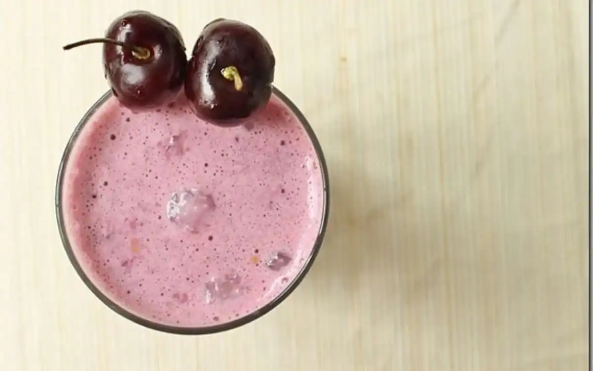 A glass of pink cherry smoothie with two whole cherries placed on the rim, viewed from above on a light wooden surface.