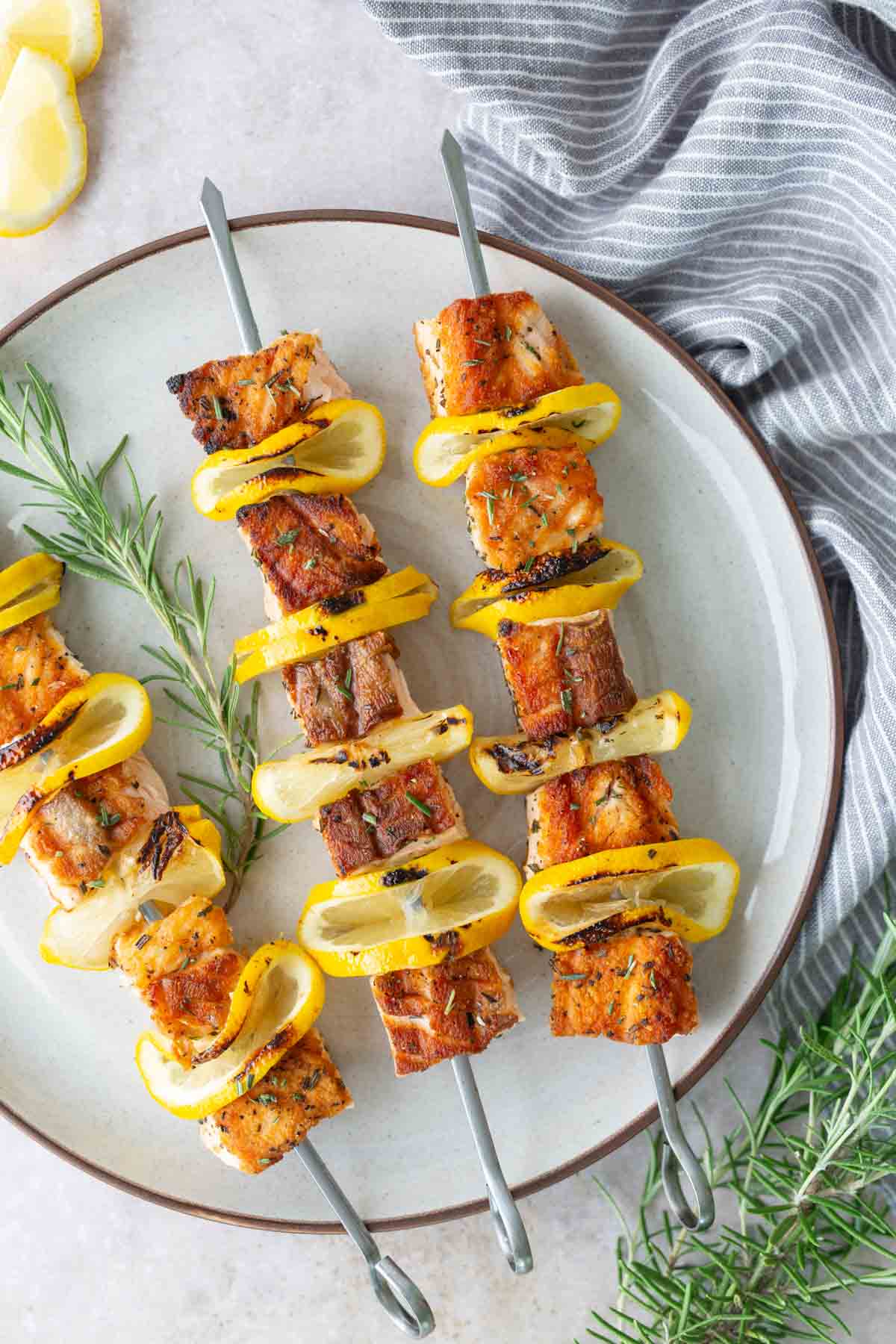 Two metal skewers with grilled fish, yellow squash slices, and lemon wedges are arranged on a white plate, garnished with fresh rosemary.