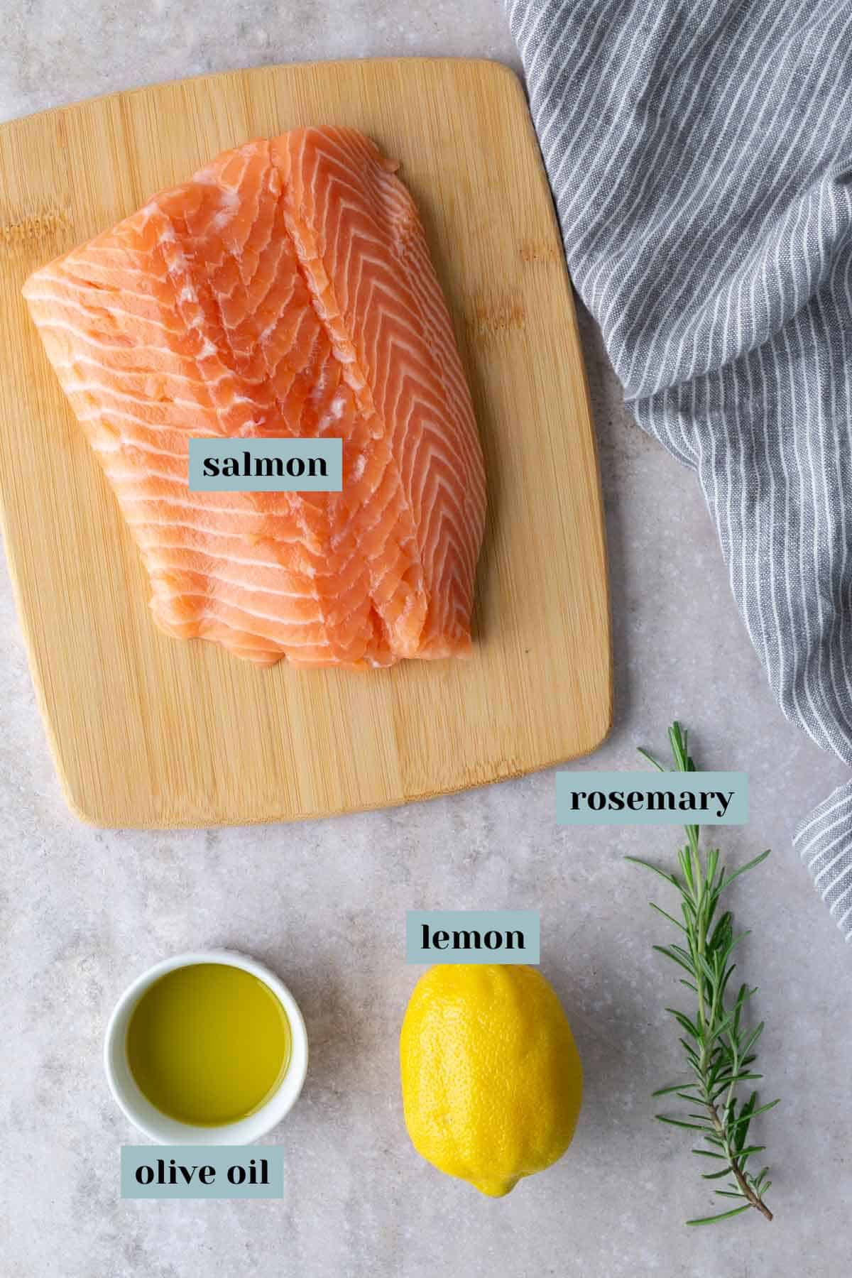 Raw salmon fillet on a wooden board, next to a sprig of rosemary, a whole lemon, and a small bowl of olive oil on a gray surface with a striped cloth.