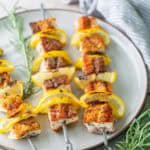 Four metal skewers with grilled fish and lemon slices are arranged on a white plate, with sprigs of rosemary beside the plate.