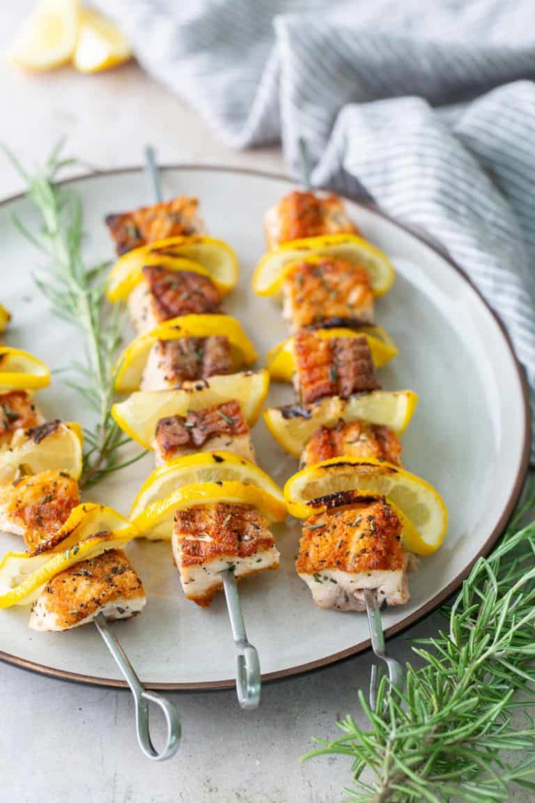 Four metal skewers with grilled fish and lemon slices are arranged on a white plate, with sprigs of rosemary beside the plate.