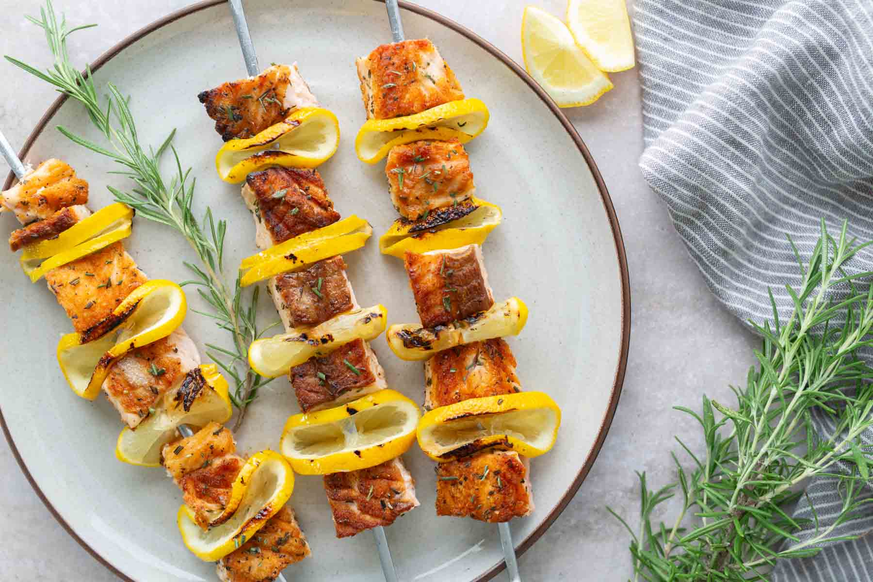 Three grilled salmon and yellow bell pepper skewers with lemon slices on a plate, garnished with fresh rosemary, with lemon wedges and a striped cloth nearby.