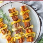 Three salmon skewers with sliced yellow squash and onions on a round plate, garnished with rosemary sprigs.