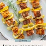 Three salmon skewers with grilled lemon slices and rosemary sprigs are arranged on a white plate. Text on the image reads "lemon rosemary salmon skewers.