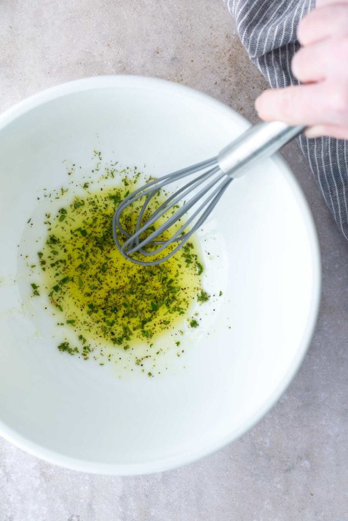 A hand holds a metal whisk mixing oil and herbs in a white bowl on a light countertop.