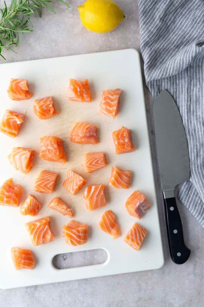 Cubed pieces of raw salmon are arranged on a white cutting board next to a knife, a sprig of rosemary, a lemon, and a gray-striped cloth.