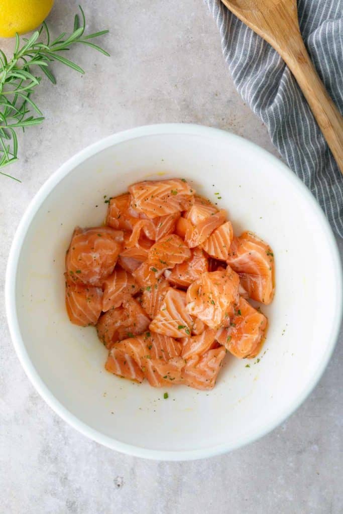 A white bowl containing raw, cubed salmon seasoned with herbs, placed on a light countertop next to a striped cloth, a wooden spoon, and fresh rosemary.