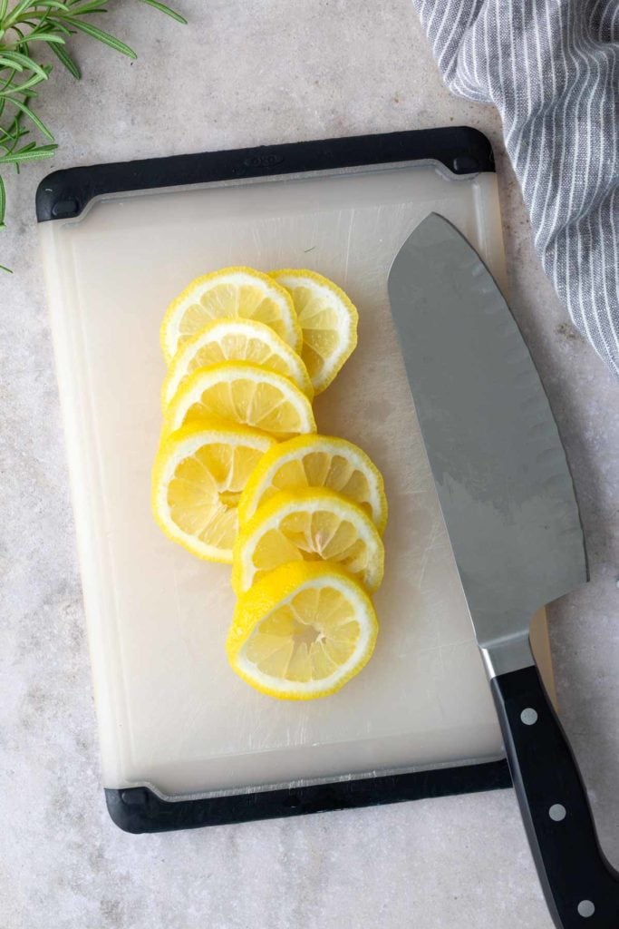 Sliced lemons arranged on a cutting board next to a kitchen knife, with a striped towel and rosemary visible nearby.