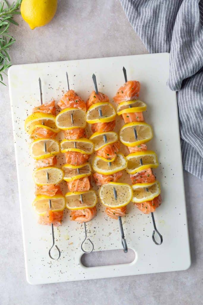 Four metal skewers with pieces of raw salmon and lemon slices are arranged on a white cutting board, seasoned with pepper. A striped towel, rosemary, and a lemon are nearby.