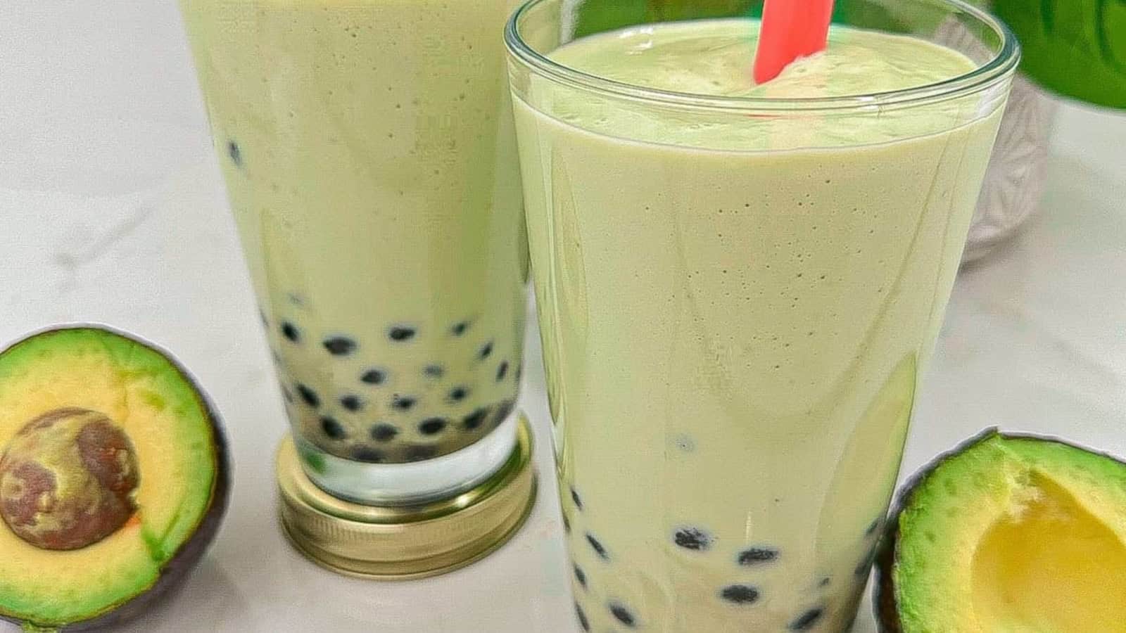 Two glasses of avocado bubble tea with black tapioca pearls, next to a halved avocado on a white surface.