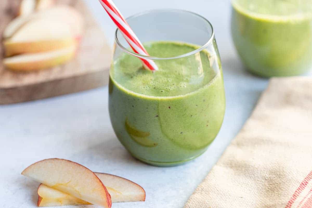 A glass of green smoothie with a red and white striped straw, next to sliced apples on a light surface.