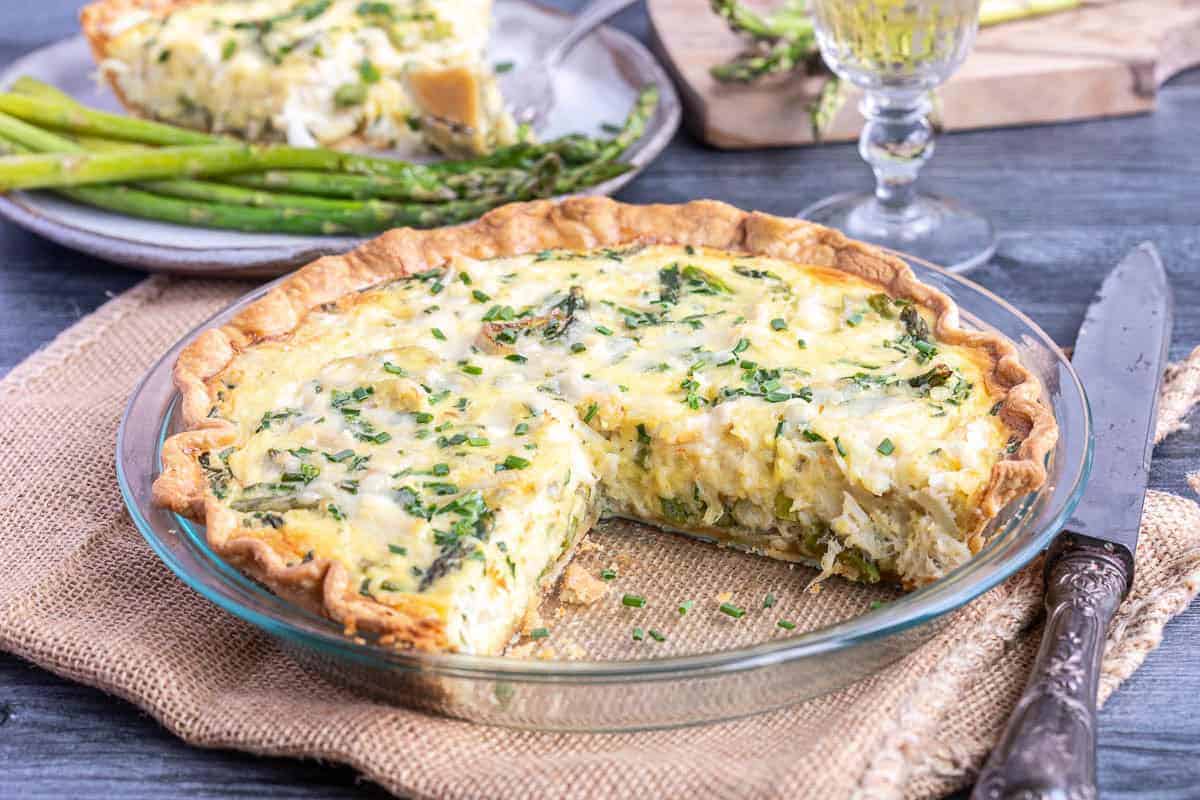 A quiche with a slice removed sits in a glass pie dish, garnished with herbs. A plate of asparagus and a glass of white wine are in the background.
