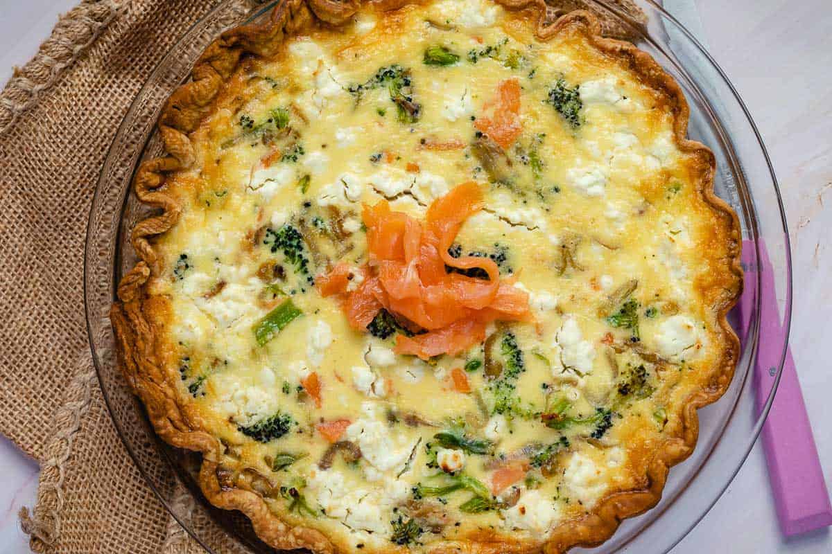 A baked quiche with broccoli, feta cheese, and smoked salmon on top, served in a glass pie dish on a rustic burlap placemat.