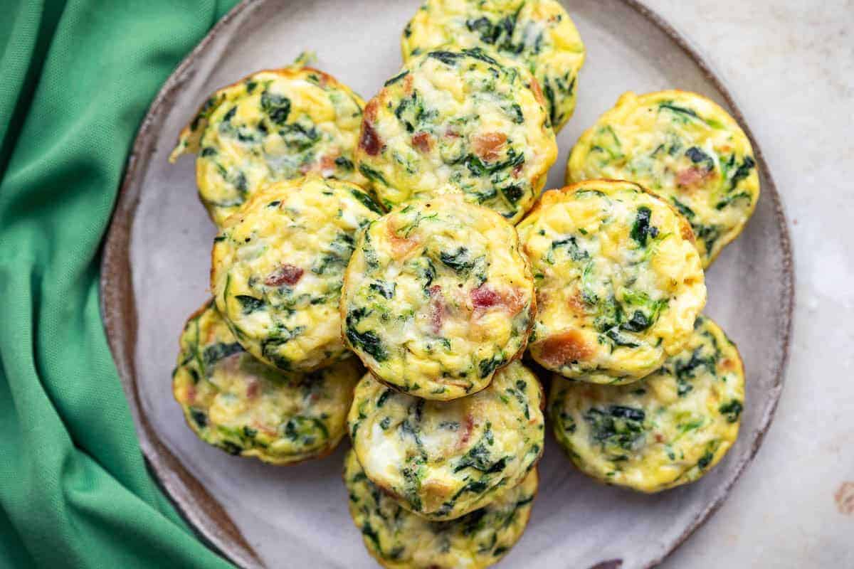 A plate of baked egg muffins with visible spinach, cheese, and bits of ham, arranged in a circular pattern next to a green cloth.