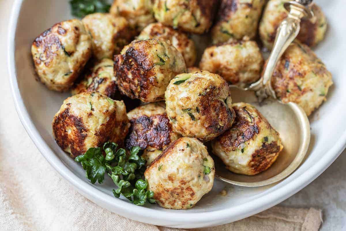 A bowl of browned meatballs garnished with parsley, with a serving spoon resting on the side.