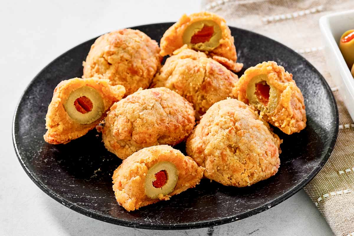 A black plate with several cheese-covered stuffed olives, some whole and some cut open to show green olives with pimentos inside.