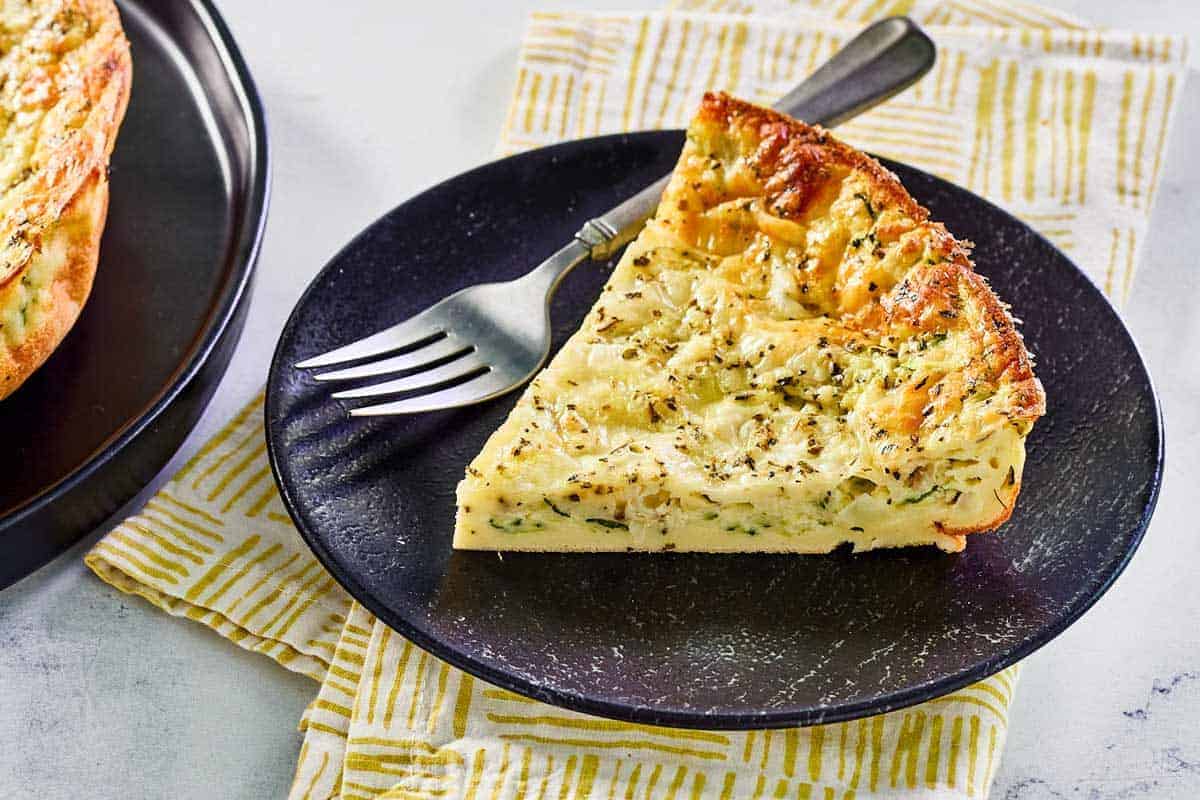 A slice of savory zucchini pie is served on a black plate with a fork, placed on a patterned yellow and white napkin.