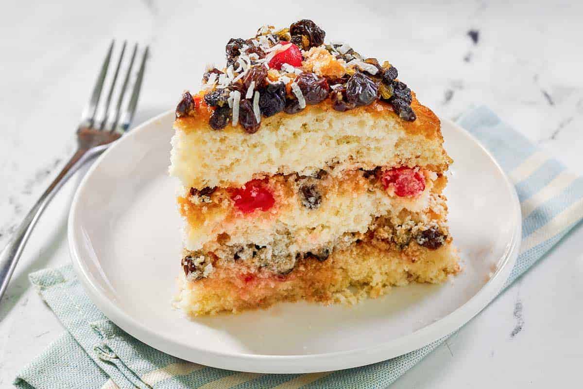 A slice of layered cake with raisins, cherries, and shredded coconut on top, served on a white plate with a fork beside it.