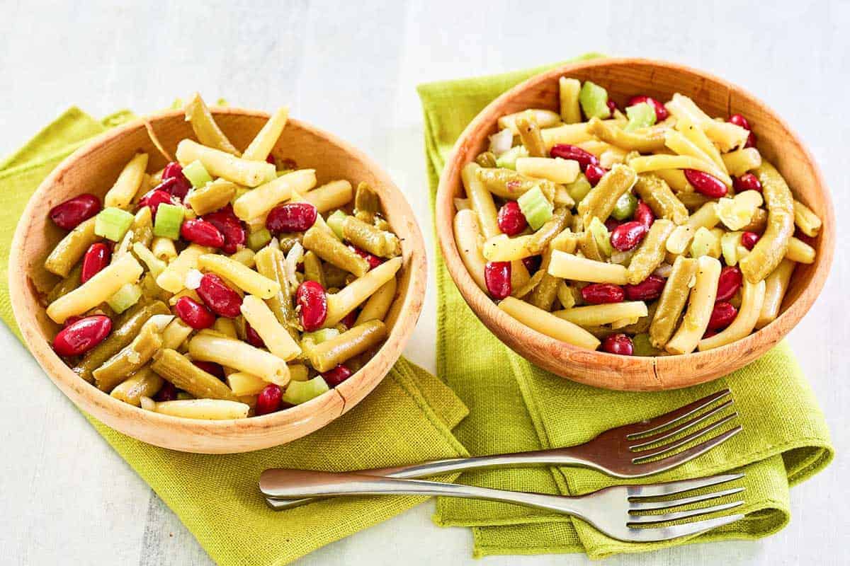 Two wooden bowls filled with three bean salad, featuring green beans, wax beans, kidney beans, and chopped celery, set on green napkins with two forks nearby.
