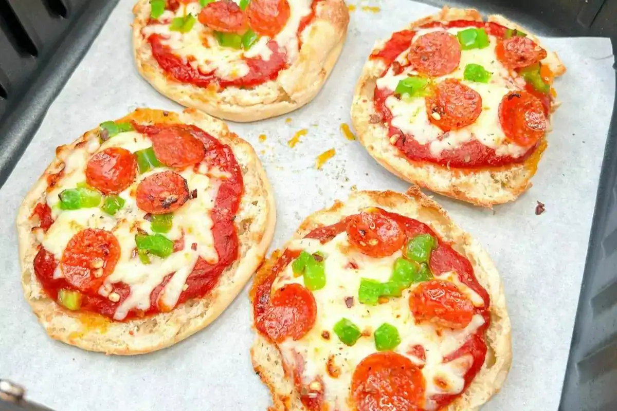Four mini pizzas with pepperoni, green bell peppers, and melted cheese are arranged on a parchment-lined baking tray.