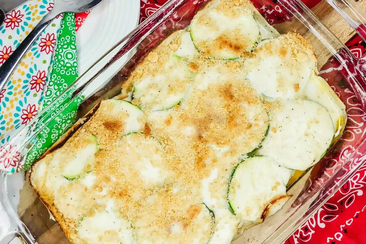 A baked casserole dish containing sliced zucchini topped with melted cheese and bread crumbs, placed on a red and green patterned cloth.