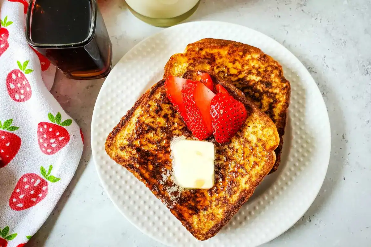 Two slices of French toast topped with a pat of butter and sliced strawberries on a white plate, with syrup and a strawberry-patterned napkin nearby.