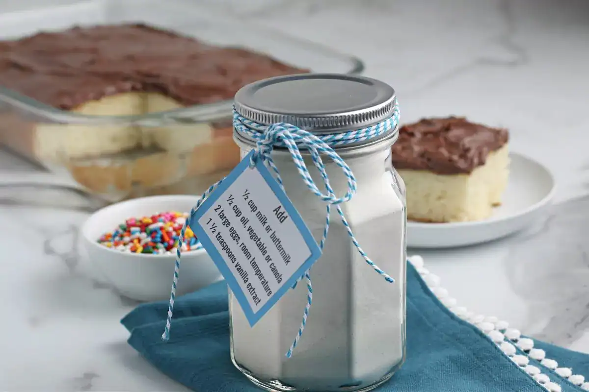 A jar of white cake mix with a recipe tag sits on a blue napkin, next to a dish of sprinkles and a frosted cake in the background.