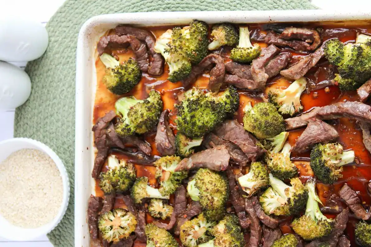 A baking dish filled with cooked beef strips and broccoli florets sits on a green mat; a bowl of sesame seeds is nearby.