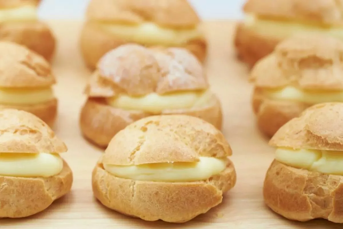 Rows of cream puffs filled with custard are arranged on a wooden surface, showcasing their golden, round pastry shells and creamy filling.