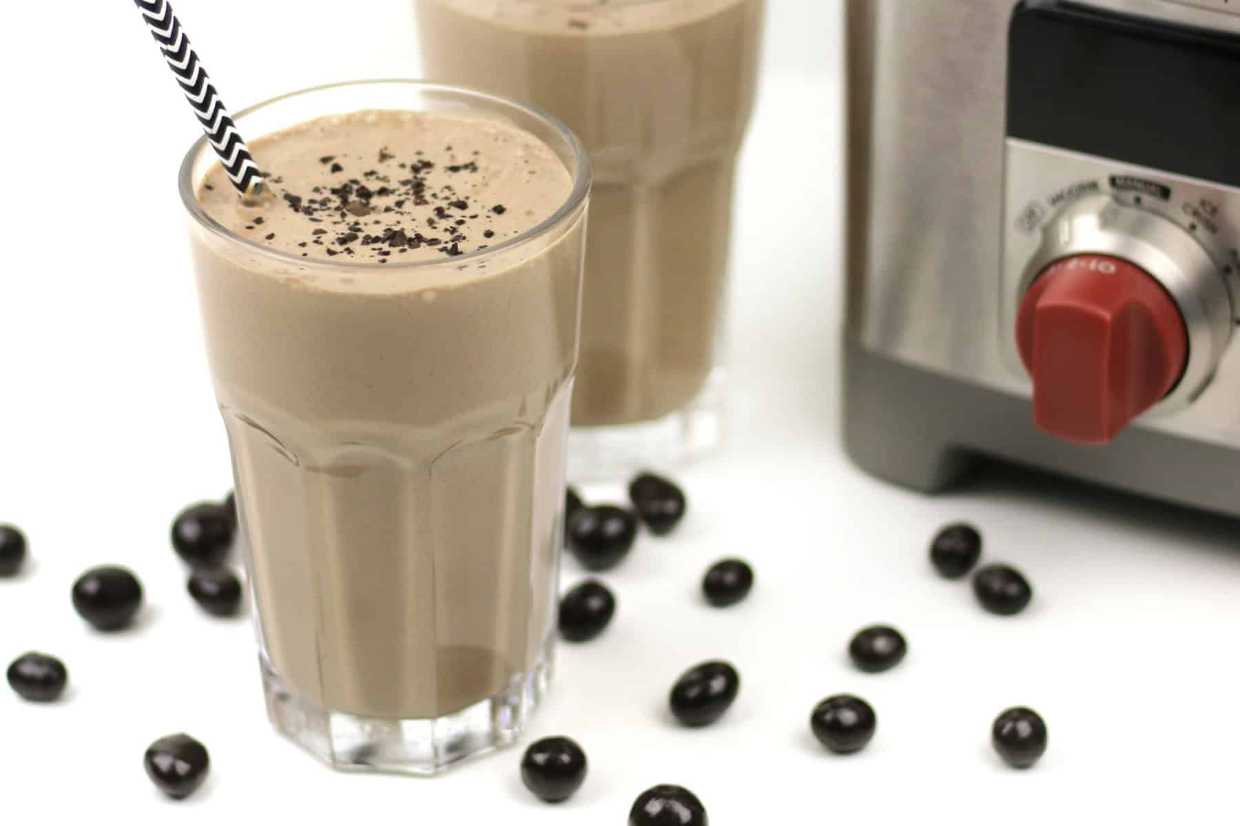 A glass of chocolate milkshake with a straw and chocolate flakes on top, next to a blender and scattered chocolate-covered coffee beans.