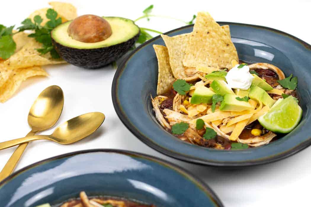 A comforting bowl of tortilla soup topped with shredded cheese, avocado, sour cream, cilantro, and lime—perfect for pantry staple recipes. Served with tortilla chips on the side, plus gold spoons and a halved avocado nearby.