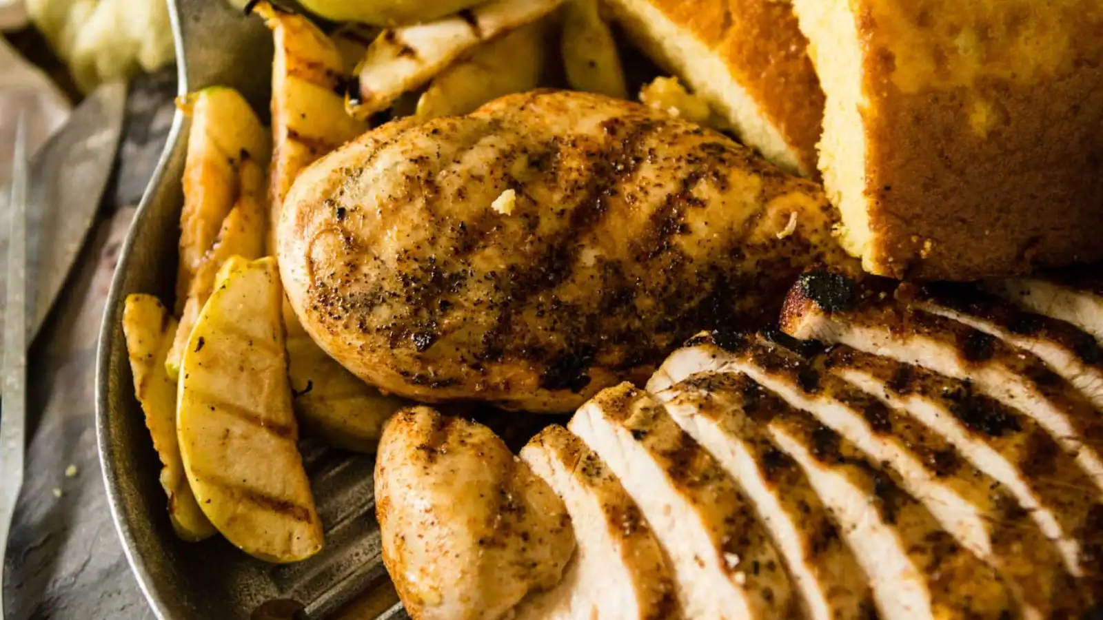 Grilled chicken breast with visible grill marks, sliced pieces, grilled vegetables, and cornbread on a metal plate.