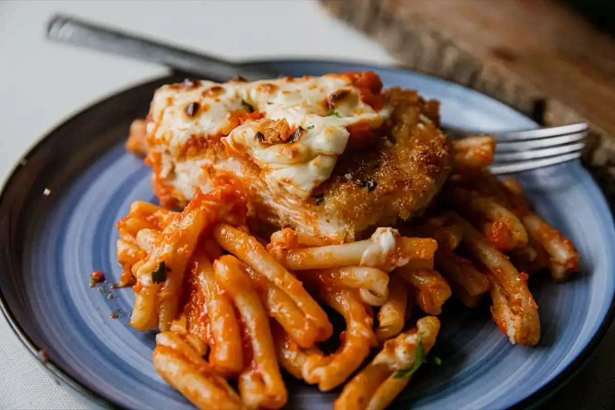 A plate of pasta with tomato sauce topped with a piece of baked chicken covered in melted cheese, with a fork on the side.