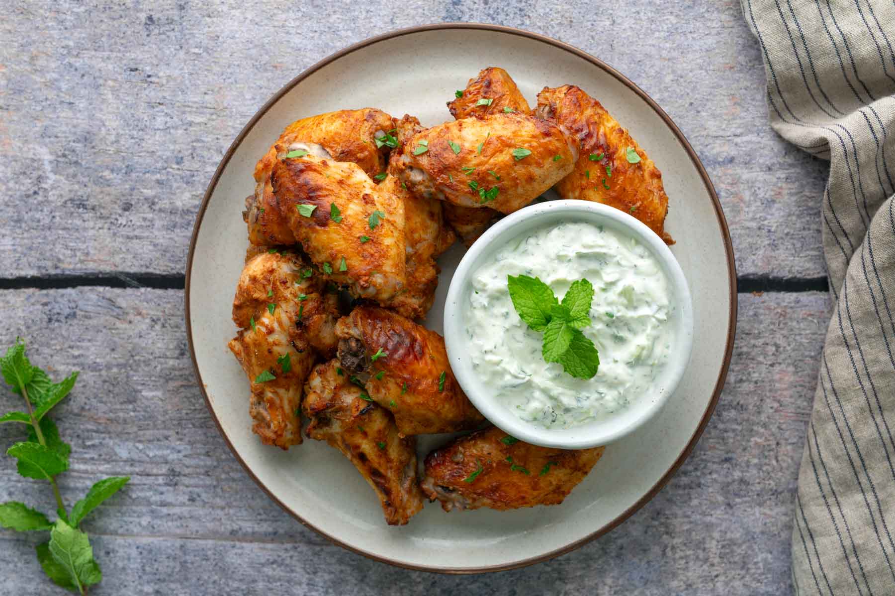 A plate of cooked chicken wings garnished with herbs, served with a bowl of creamy dipping sauce topped with a mint sprig, on a stone surface next to a striped cloth.